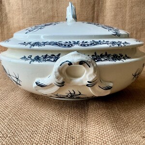 Puede incluir: Una fuente de cer&aacute;mica blanca y azul con tapa. La fuente presenta un patr&oacute;n floral en azul y tiene asas a ambos lados. La tapa tiene un adorno decorativo. La fuente es probablemente vintage o antigua.