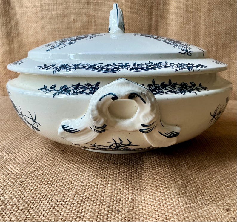 Puede incluir: Una fuente de cer&aacute;mica blanca y azul con tapa. La fuente presenta un patr&oacute;n floral en azul y tiene asas a ambos lados. La tapa tiene un adorno decorativo. La fuente es probablemente vintage o antigua.