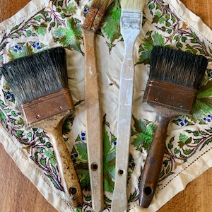 Op de afbeelding: Vier vintage penselen met houten handvatten en koperen bussen liggen op een bloemen geborduurde doek. De penselen vertonen tekenen van slijtage. De doek heeft groene, paarse en blauwe bloempatronen. De penselen variëren in grootte en haarkleur.