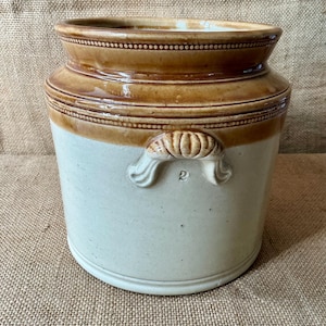 May include: A cylindrical stoneware crock featuring a cream-coloured body and a glossy brown glazed rim. The vessel includes two moulded lug handles on the sides, with the number 2 embossed below one handle, and a decorative beaded band detail.