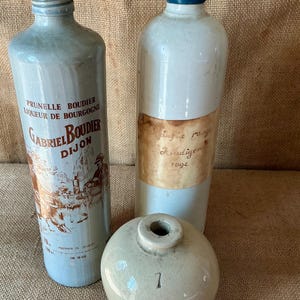 May include: Two tall ceramic bottles and one short, round ceramic vessel. The left bottle is light blue with a label reading Prunelle Boudier Liqueur de Bourgogne Gabriel Boudier Dijon. The right bottle has a blue cap and a tan label. The round vessel has a 7.