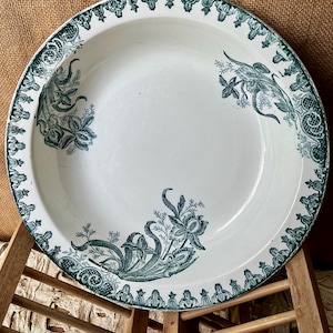 Könnte beinhalten: Weißer Keramik-Servierteller mit breitem Rand und flacher Schüssel. Der Rand und das Innere des Tellers sind mit einem teal-farbenen Blumenmuster verziert. Das Blumenmuster umfasst stilisierte Blumen und Blätter.