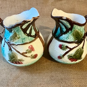 Peut inclure: Deux pichets en céramique avec un motif floral. Les pichets ont une base blanche avec des accents marron, sarcelle et rouge. Chaque pichet a une anse et un bord marron. Les pichets mesurent environ 13 cm de haut.