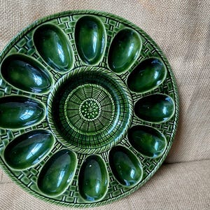 May include: A round, dark green ceramic egg serving platter featuring twelve oval-shaped indentations arranged in a circle around a central circular compartment. The surface has a raised, textured basket-weave pattern throughout the entire plate.
