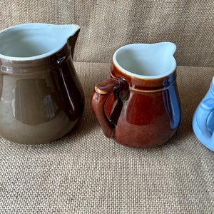 Może przedstawiać: Trzy ceramiczne dzbanki o różnych rozmiarach i kolorach. Największy jest błyszczący brązowy, średni jest ciemnoczerwono-brązowy, a najmniejszy jest jasnoniebieski. Każdy dzbanek ma zakrzywiony uchwyt, szeroki dzióbek i gładkie białe wnętrze.