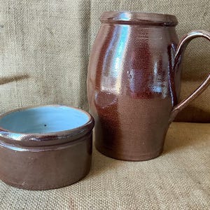 Op de afbeelding: Een bruin geglazuurde keramische kan met een gebogen handvat en een bijpassende lage kom met een gladde lichtblauwe binnenkant. Beide stukken hebben een glanzende afwerking en een ronde vorm met een consistente donkerbruine buitenkant.