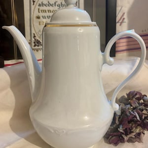 Peut inclure: Une cafetière en porcelaine blanche avec un bec verseur et une anse incurvés. Le pot a une bordure dorée autour du couvercle et un bouton doré. La base du pot présente un motif en relief subtil. En arrière-plan, un point de croix.