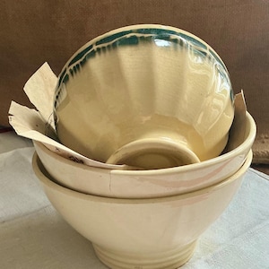 Op de afbeelding: Een stapel van drie crèmekleurige keramische kommen. De bovenste kom heeft een geschulpte rand met een teal groene rand. De kommen zijn genest, met de grootste kom onderaan en een kleinere kom erin.