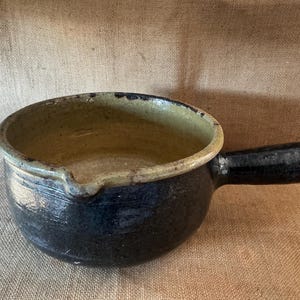 Puede incluir: Un cazo de cerámica redondo con un exterior negro oscuro y un interior esmaltado en tono bronceado claro. El recipiente cuenta con un mango único y resistente en el lateral y un pequeño pico vertedor integrado en el borde superior.