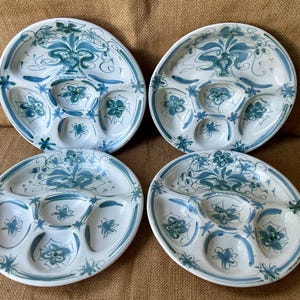 Puede incluir: Un juego de cuatro platos divididos de cerámica blanca redonda, cada uno con un patrón floral pintado a mano en azul y verde azulado. Los platos están segmentados en cinco secciones distintas, incluyendo un compartimento central circular.