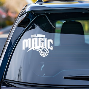 May include: White vinyl decal of the Orlando Magic NBA team logo on the back window of a black car. The logo features the team name "Orlando Magic" with a basketball and a shooting star.