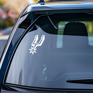 Może przedstawiać: Biała naklejka z logo San Antonio Spurs na tylnej szybie czarnego samochodu.