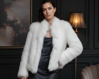 May include: A white faux fur jacket with a wide collar. The jacket is worn over a dark gray satin dress. The background features a dark wall, a framed painting, and a floor lamp.