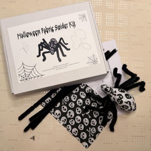 May include: A Halloween Fabric Spider Kit in a white box, featuring a black spider illustration and the text "Halloween Fabric Spider Kit". Includes fabric, instructions, and a spider with black pipe cleaner legs and a skull-patterned body.
