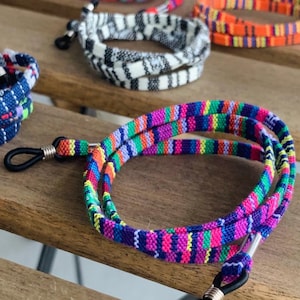 May include: A colourful, striped eyeglass chain made of woven fabric. The chain has a silver metal clasp and black rubber tips.