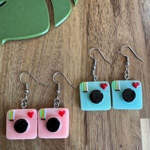 May include: Earrings with a square camera design in pink and light blue. Each earring has a black lens, a red heart, and colourful stripes. The earrings hang from silver-coloured hooks.