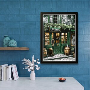 a framed painting of a pub with a green door and windows, a Guinness sign, and barrels outside, hanging on a blue tiled wall above a white table with a vase of flowers and a bowl of fruit.