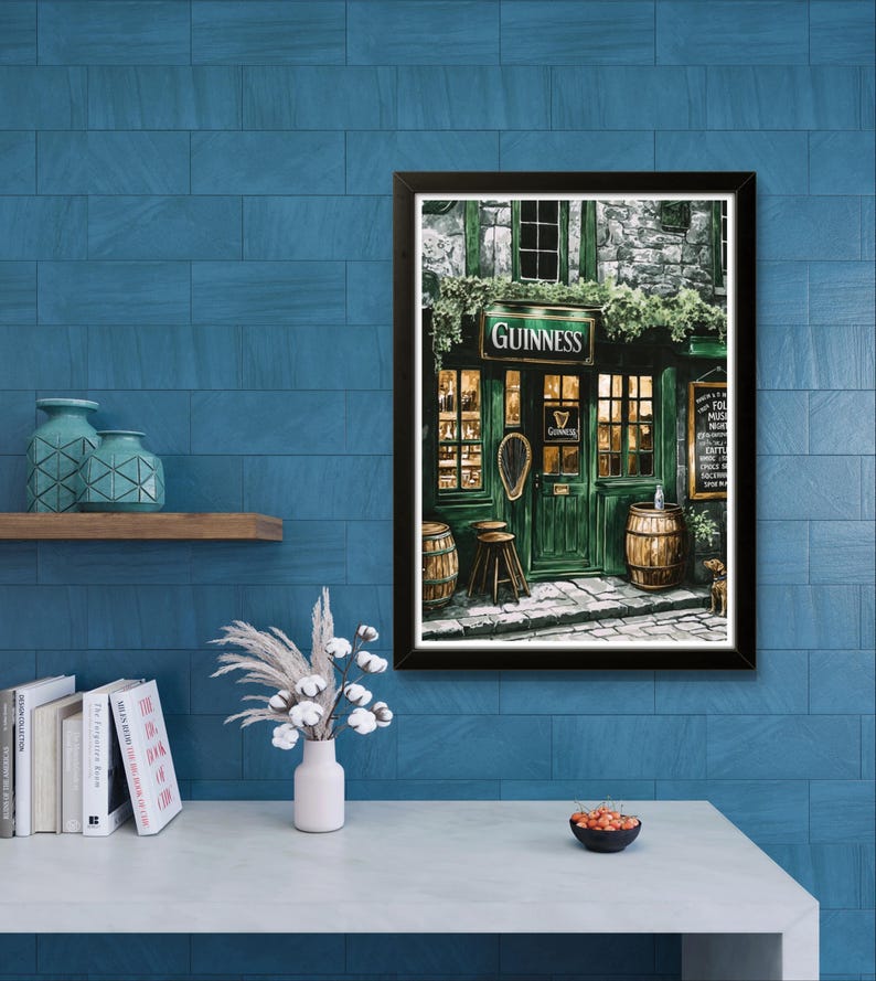 a framed painting of a pub with a green door and windows, a Guinness sign, and barrels outside, hanging on a blue tiled wall above a white table with a vase of flowers and a bowl of fruit.