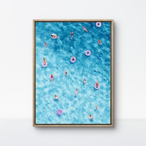 Puede incluir: Una impresión artística enmarcada que representa una vibrante masa de agua azul con numerosas figuras nadando y flotando en flotadores inflables rosas. La obra de arte tiene un estilo texturizado y pictórico, que evoca un día soleado en la playa o la piscina.