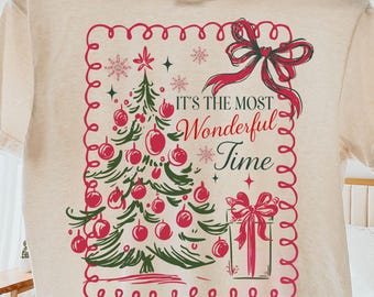 Op de afbeelding: Crèmekleurig T-shirt met een kerstboomontwerp met rode ballen en een rode strik. De tekst "It's the Most Wonderful Time" is weergegeven in een feestelijk lettertype, perfect voor de feestdagen.
