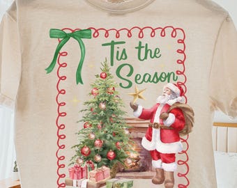 May include: Cream-colored t-shirt featuring a Christmas-themed graphic. The design includes a decorated Christmas tree, Santa Claus, and the text "Tis the Season" in a red and green frame. A green bow is at the top left.