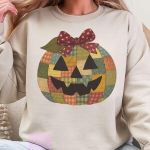 May include: Beige sweatshirt featuring a patchwork pumpkin design. The pumpkin has a smiling face, a green leaf, and a burgundy bow. The pumpkin is made of various colors, including green, orange, yellow, and red. A cozy fall-themed garment.