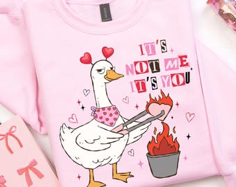 May include: A light pink sweatshirt with a cartoon goose design. The goose is wearing a bandana and holding tongs over a heart-shaped flame. Text on the shirt reads "IT'S NOT ME IT'S YOU". Decorative hearts and stars surround the goose.