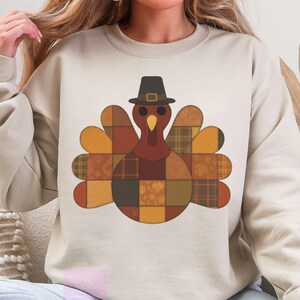 May include: Beige crewneck sweatshirt featuring a patchwork turkey design. The turkey has a brown body, a red head, and a black pilgrim hat. The wings are in shades of orange and yellow. A festive and cozy apparel item for Thanksgiving.