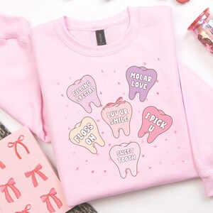 K&ouml;nnte beinhalten: Hellrosa Sweatshirt mit Cartoon-Zahngrafiken und Spr&uuml;chen wie "Filling Special" und "Sweet Tooth". Das Bild enth&auml;lt dekorative Elemente wie Makronen und einen Becher, was eine verspielte &Auml;sthetik erzeugt.