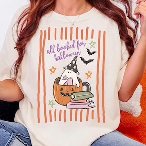 May include: Cream-colored t-shirt with a Halloween-themed graphic. The design features a ghost wearing a witch hat inside a pumpkin mug, with the text "all booked for halloween". Includes stars and bats.