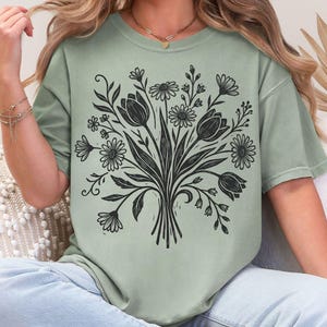 May include: Sage green t-shirt featuring a black floral bouquet design. The graphic includes tulips, daisies, and other flowers. The shirt has a relaxed fit and short sleeves.
