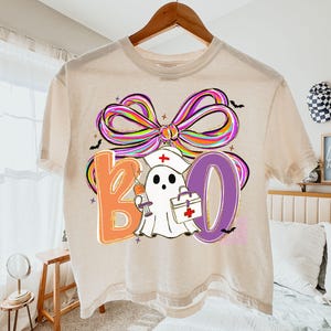 Pu&ograve; includere: Una maglietta beige con un design grafico colorato. Il design presenta la parola "BOO" in arancione e viola, un fantasma con un cappello da infermiera e un fiocco arcobaleno. Il fantasma tiene in mano una siringa e una borsa medica.