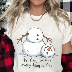 May include: A cream-colored t-shirt featuring a graphic of a large snowman with a smaller, detached snowman head resting beside it. The design includes blue snowflakes and the text "it's fine, i'm fine everything is fine" in black lettering.