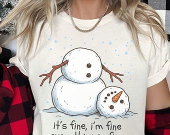May include: A cream-colored t-shirt featuring a graphic of a large snowman standing next to a smaller, fallen snowman head. The design includes blue snowflakes and the text "it's fine, i'm fine everything is fine" printed in black lettering.