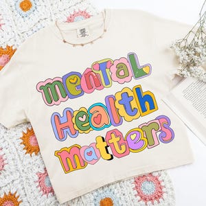 May include: Cream-colored t-shirt with the words "Mental Health Matters" in colorful, bubble letters. The shirt is laid on a crocheted blanket with floral patterns. A necklace and a book are also present.