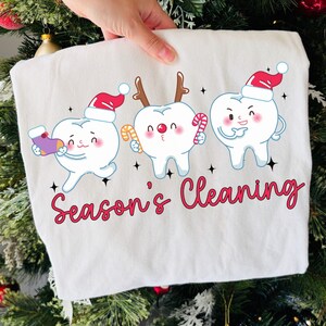 Puede incluir: Una sudadera blanca con tres dientes de dibujos animados con accesorios navide&ntilde;os. Los dientes llevan gorros de Pap&aacute; Noel y cuernos de reno, sosteniendo bastones de caramelo. Las palabras "Season's Cleaning" est&aacute;n escritas en rojo.