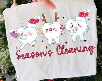 May include: A white sweatshirt featuring three cartoon teeth with festive holiday accessories. The teeth are wearing Santa hats and reindeer antlers, holding candy canes and a stocking. The words "Season's Cleaning" are written in red script.