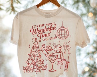 May include: Cream-colored t-shirt with a red graphic. The graphic features a Christmas tree, Santa Claus in a martini glass, and the text "IT'S THE MOST Wonderful Time OF THE YEAR". A disco ball and champagne glasses are also included.