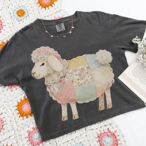 Könnte beinhalten: Dunkelgraues T-Shirt mit einem Patchwork-Schaf-Motiv. Das Schaf hat ein cremefarbenes Gesicht und Ohren sowie einen Körper aus floralen und rosa gemusterten Flicken. Eine goldene Schmetterlingskette ist sichtbar. Das Shirt liegt auf einer weißen Häkeldecke.
