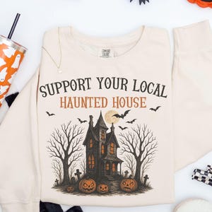 May include: A cream-colored crewneck sweatshirt featuring a graphic print with the text SUPPORT YOUR LOCAL HAUNTED HOUSE in black and orange. The design includes a dark house, bare trees, bats, a full moon, and orange jack-o'-lantern pumpkins.