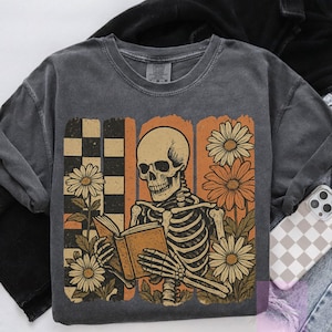 May include: Dark gray sweatshirt featuring a skeleton reading a book, surrounded by daisies. The design includes a checkered pattern and orange vertical stripes. The skeleton and flowers are in shades of beige, orange, and white.