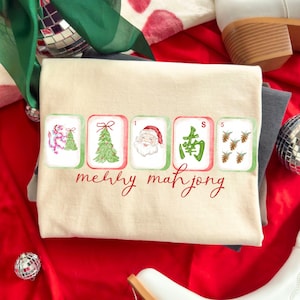 May include: A cream-colored t-shirt featuring a graphic of five mahjong tiles arranged horizontally. The tiles display a dragon, a Christmas tree, Santa Claus, a green Chinese character, and pinecones. The text "merry mahjong" is printed below.