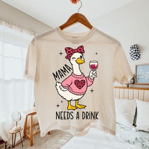 Peut inclure: T-shirt cr&egrave;me avec un dessin d'oie de dessin anim&eacute; portant un pull rose avec un motif de c&oelig;ur et un n&oelig;ud. L'oie tient un verre de vin rouge. Le texte sur le t-shirt se lit "MAMA NEEDS A DRINK".