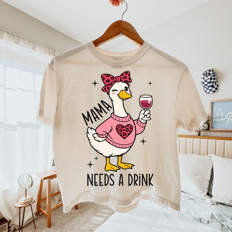 Peut inclure: T-shirt cr&egrave;me avec un dessin d'oie de dessin anim&eacute; portant un pull rose avec un motif de c&oelig;ur et un n&oelig;ud. L'oie tient un verre de vin rouge. Le texte sur le t-shirt se lit "MAMA NEEDS A DRINK".