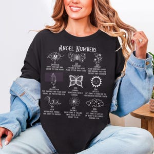 以下が含まれることがあります： 「Angel Numbers」の文字と様々なシンボルと数字がプリントされた黒の長袖シャツ。目、蝶、花、その他のスピリチュアルなシンボルのイラストが特徴です。「自分の直感を信じて」などのフレーズが含まれています。