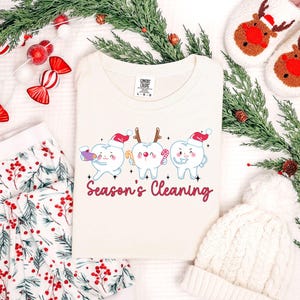 Puede incluir: Camiseta color crema con un dise&ntilde;o de tres dientes de dibujos animados con gorros de Pap&aacute; Noel y bastones de caramelo. El texto "Season's Cleaning" est&aacute; en escritura roja. Decoraciones navide&ntilde;as en el fondo.