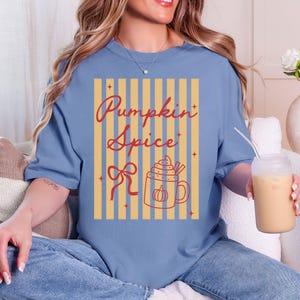 May include: A light blue t-shirt with a fall-themed graphic. The graphic features the words "Pumpkin Spice" in a cursive font, a bow, and a mug of pumpkin spice latte. The shirt has a relaxed fit.