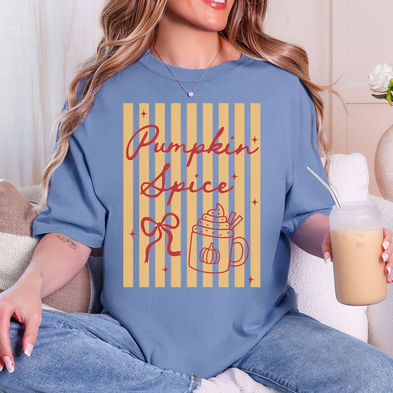 May include: A light blue t-shirt with a fall-themed graphic. The graphic features the words "Pumpkin Spice" in a cursive font, a bow, and a mug of pumpkin spice latte. The shirt has a relaxed fit.