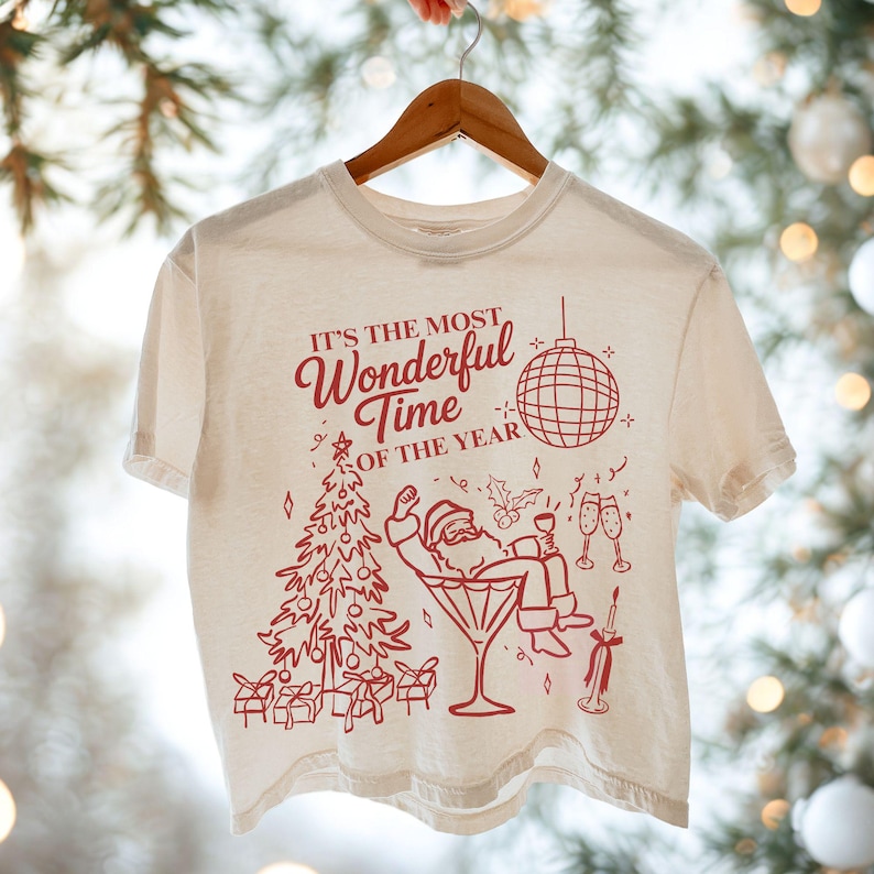 Puede incluir: Camiseta color crema con un gr&aacute;fico rojo navide&ntilde;o. El dise&ntilde;o incluye un &aacute;rbol de Navidad, Pap&aacute; Noel en una copa de Martini y el texto "IT'S THE MOST Wonderful Time OF THE YEAR". Tambi&eacute;n se muestra una bola de discoteca y copas de champ&aacute;n.