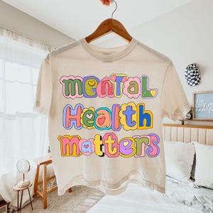 May include: A cream-colored t-shirt with the words "Mental Health Matters" in colorful, bubble letters. The text is outlined in various colors, including pink, blue, yellow, and green, with heart accents. The shirt is hanging on a wooden hanger.